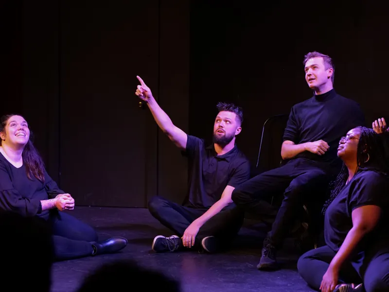 Four performers seated on the floor in magenta side light, one pointing upward