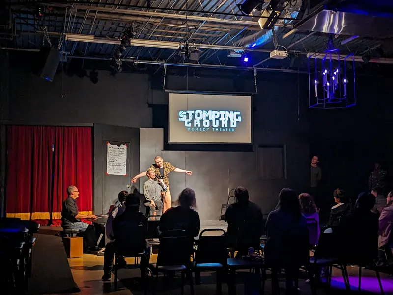 Full house during a show, warm stage light on the performers and the Stomping Ground screen above