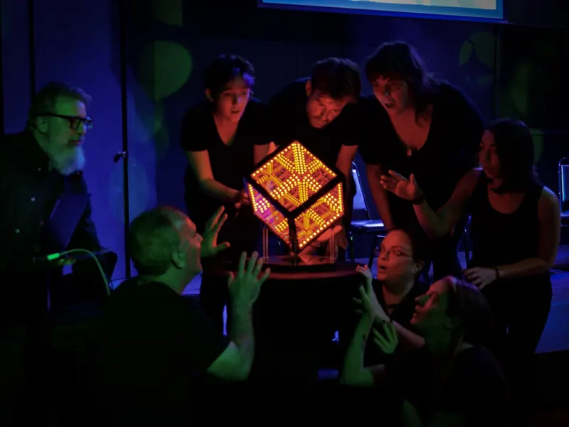 Performers gathered around a glowing orange cube prop under green and blue light