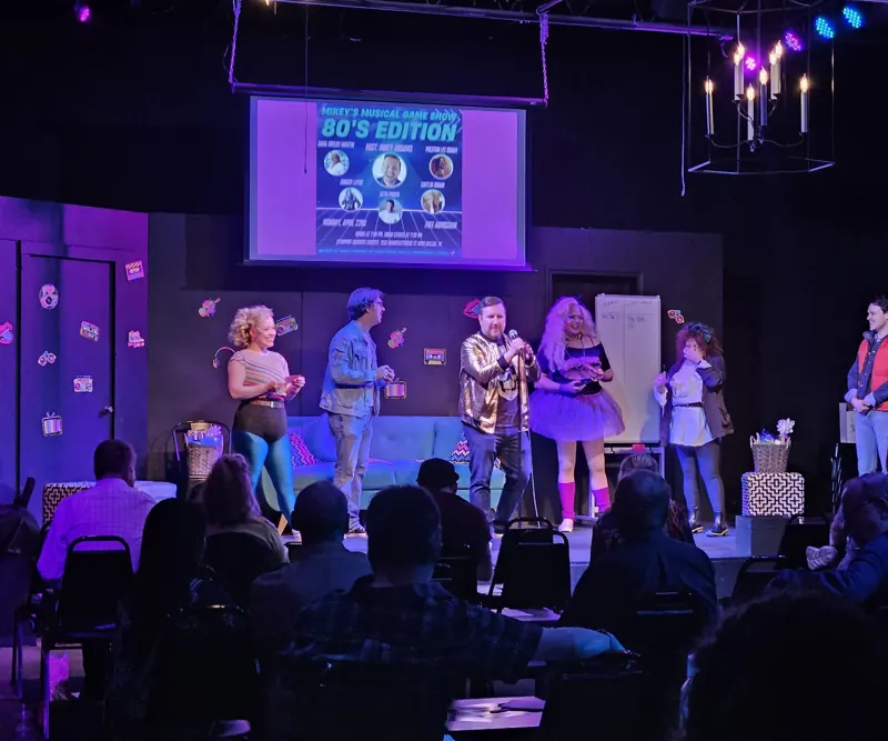 Ensemble across the stage in purple and amber for an 80s-themed night, crowd in the foreground