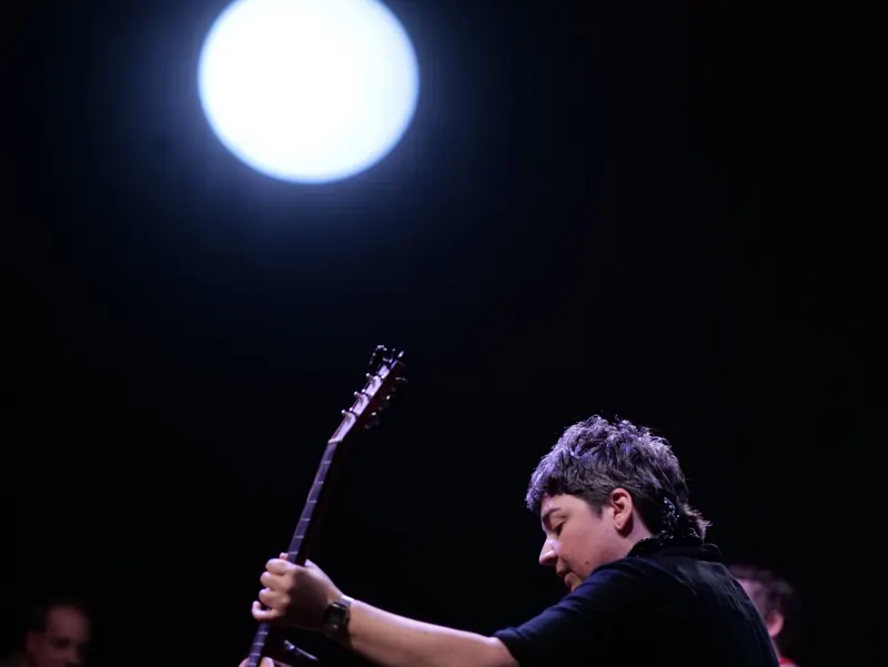 Guitarist lit from the side against black, with a single bright moon-like spot behind